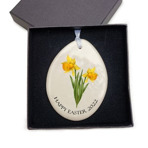 Pode incluir: Ornamento de ovo de P&aacute;scoa de cer&acirc;mica branca com um design de flores de narciso amarelo e o texto "Happy Easter 2022".