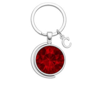 May include: A silver-colored keychain with a round, faceted, deep red pendant. A small silver letter "C" charm hangs next to the pendant. The keychain ring is also silver.