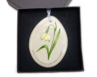 May include: An egg-shaped ceramic ornament with a painted snowdrop flower design, presented in a dark gray box. The ornament has a light blue ribbon for hanging and features the text "HAPPY EASTER 2022 LOVE GARRY, MELISSA & FAMILY."