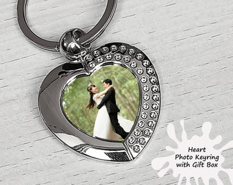 May include: Silver heart-shaped photo keychain with a picture of a couple in a wedding setting. The keychain has a small ring for attaching keys. The text "Heart Photo Keyring with Gift Box" is printed on a white splatter background.