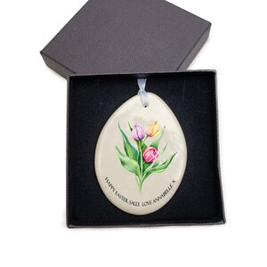May include: A white ceramic egg-shaped ornament with a painted image of three tulips in shades of pink, yellow, and purple. The ornament has a ribbon attached and the text "Happy Easter Sally Love Annabelle x" is written around the bottom.