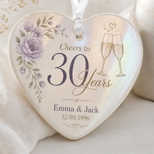 May include: A heart-shaped ceramic ornament with an iridescent finish, featuring a purple floral illustration, two champagne flutes, and the text Cheers to 30 Years, Emma & Jack, and 12.08.1996. A white ribbon is attached to the top for hanging.