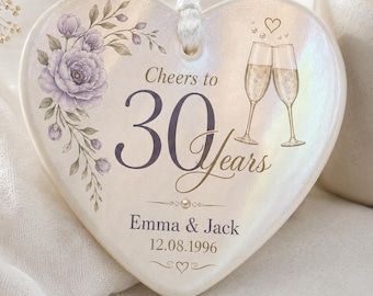 May include: A heart-shaped ceramic ornament with an iridescent finish, featuring a purple floral illustration, two champagne flutes, and the text Cheers to 30 Years, Emma & Jack, and 12.08.1996. A white ribbon is attached to the top for hanging.