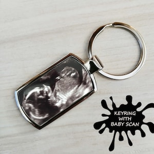 May include: Silver keychain with a black and white ultrasound image of a baby inside a rectangular frame. The text "KEYRING WITH BABY SCAN" is printed on a black splatter design.