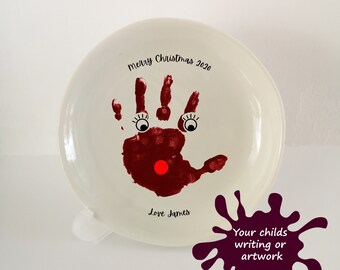 May include: White ceramic plate with a red handprint, black eyes, and a red nose. The plate has the text "Merry Christmas 2020" and "Love James" written on it.