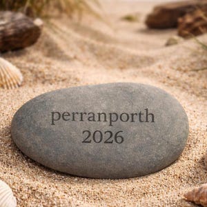 May include: A gray, oval stone with the inscription "perranporth 2026" engraved on its surface. The stone rests on a sandy beach, surrounded by seashells and beach grass. The ocean is visible in the background.