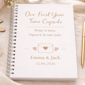 May include: A white, spiral-bound journal titled "Our First Year Time Capsule." The journal includes the text "Write it now. Open it in one year." and the names "Emma & Jack" with a date. A gold pen rests beside the journal.