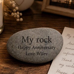 May include: A gray engraved stone with the words "My rock", "Happy Anniversary", and "Love Wifey". A candle, a decorative heart, and a framed photo are in the background. A handwritten note is also visible.