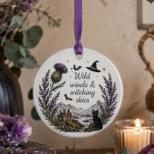 May include: A round, white ceramic ornament with a purple ribbon for hanging. The ornament features a black cat, a witch's hat, and the text "Wild winds & witching skies." The design includes a thistle, mountains, and a castle.