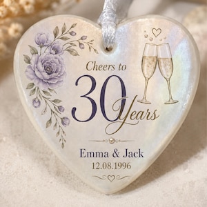 May include: A heart-shaped ceramic ornament with an iridescent finish, featuring a purple floral illustration, two champagne flutes, and the text "Cheers to 30 Years," "Emma & Jack," and "12.08.1996." A white ribbon is attached to the top for hanging.