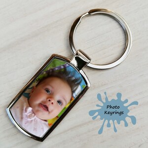 A silver rectangular metal keyring displaying a close-up photo of a smiling baby with curly hair. The keyring is lying on a pale wooden background next to a blue paint splash graphic that says "Photo Keyrings."