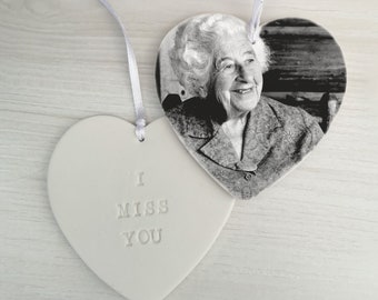 May include: A white ceramic heart ornament with the text "I Miss You" on one side and a black and white photo of an older woman on the other side.