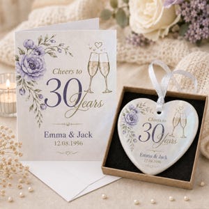 May include: A white greeting card and a heart-shaped ceramic ornament in a box, both featuring purple floral illustrations, two champagne flutes, and the text Cheers to 30 Years, Emma & Jack, 12.08.1996. The ornament has a white ribbon for hanging.