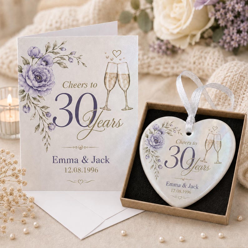 May include: A white greeting card and a heart-shaped ceramic ornament in a box, both featuring purple floral illustrations, two champagne flutes, and the text Cheers to 30 Years, Emma & Jack, 12.08.1996. The ornament has a white ribbon for hanging.