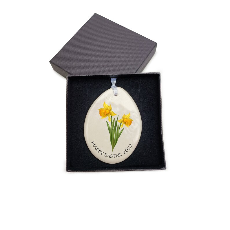 Pode incluir: Ornamento de ovo de P&aacute;scoa de cer&acirc;mica branca com um design de flores de narciso amarelo e o texto "Happy Easter 2022".