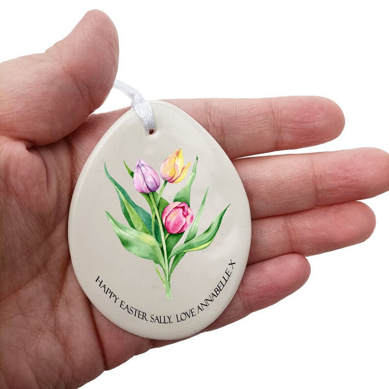 May include: A white ceramic egg-shaped ornament with a watercolor painting of pink, yellow, and purple tulips. The ornament has a white ribbon attached and the text "Happy Easter Sally. Love Annabelle x" is printed on the bottom.