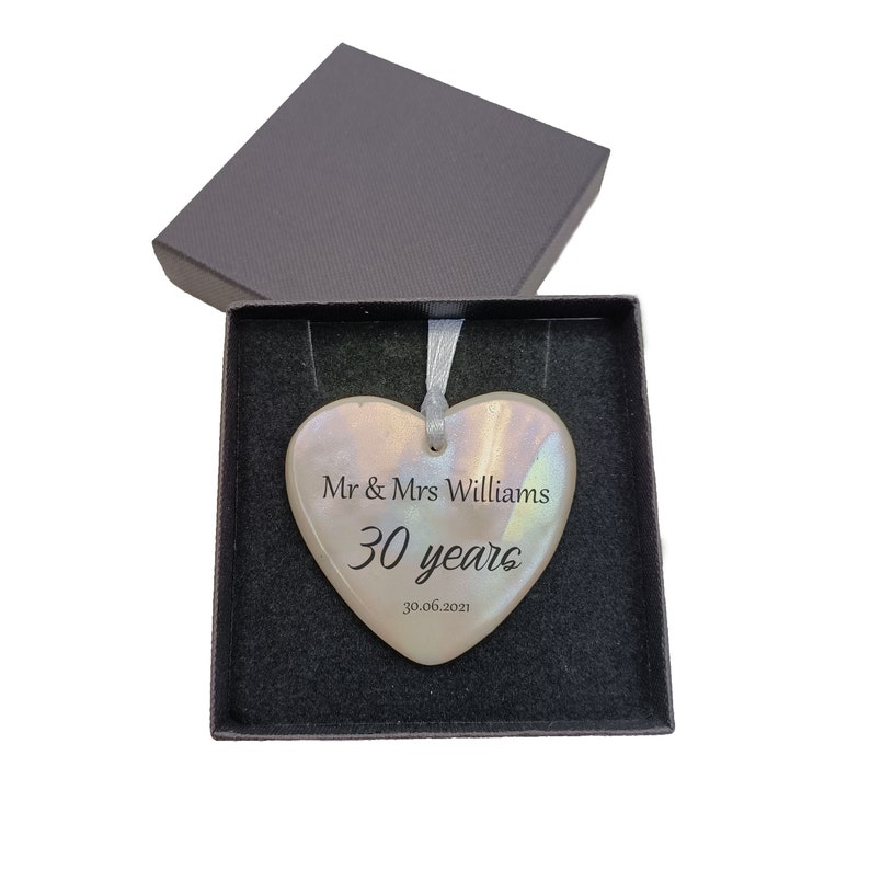 May include: A heart-shaped ceramic ornament in a black gift box. The ornament is decorated with a light brown glaze and has a white ribbon attached. The ornament is personalized with the text "Mr & Mrs Williams 30 years 30.06.2021".