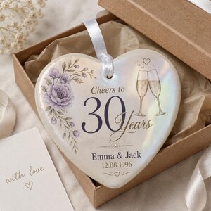 May include: A heart-shaped ceramic ornament with an iridescent finish, featuring a purple floral illustration, two champagne flutes, and the text Cheers to 30 Years, Emma & Jack, and 12.08.1996. A white ribbon is attached for hanging.