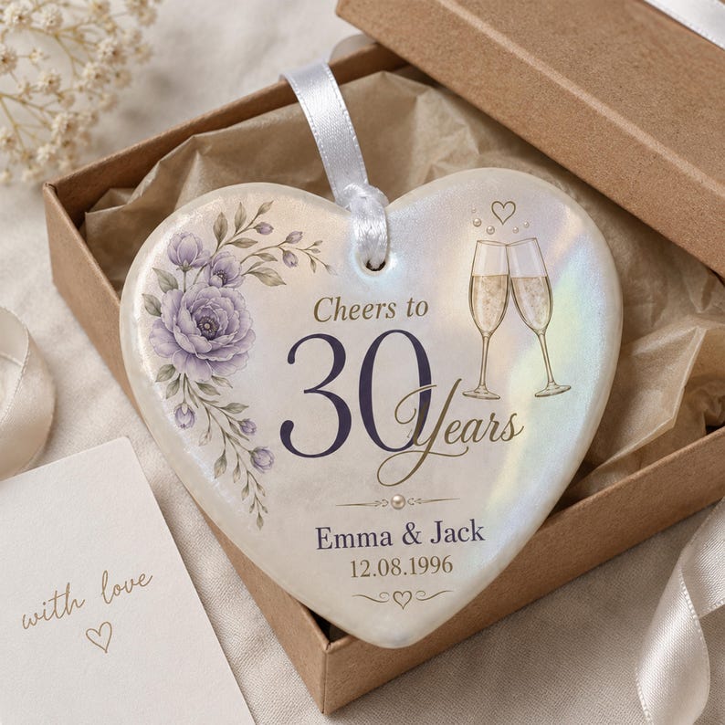 May include: A heart-shaped ceramic ornament with an iridescent finish, featuring a purple floral illustration, two champagne flutes, and the text Cheers to 30 Years, Emma & Jack, and 12.08.1996. A white ribbon is attached for hanging.