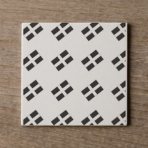 May include: Square ceramic tile with a repeating pattern of the Cornish flag, a white cross on a black background. The tile has a smooth, matte finish and is set against a wooden surface. The design is simple and graphic.