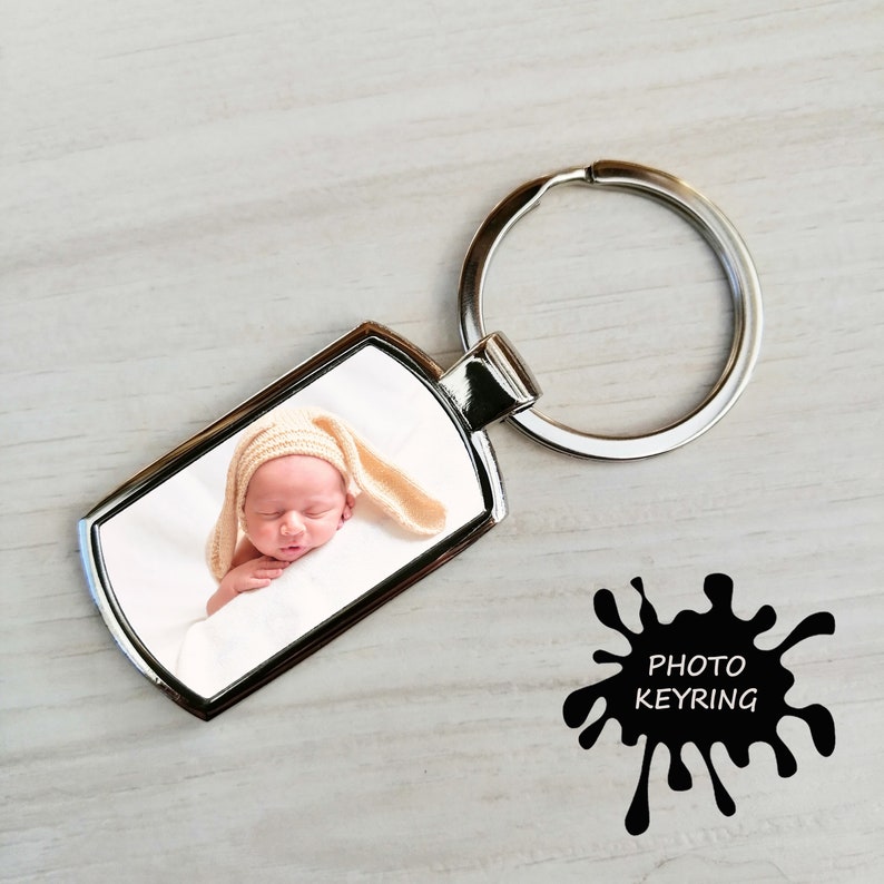 A silver metal keyring featuring a photo of a sleeping newborn baby wearing a soft beige knitted hat with long floppy ears. The keyring lies on a light wood surface with a black paint splatter graphic labeled "PHOTO KEYRING."
