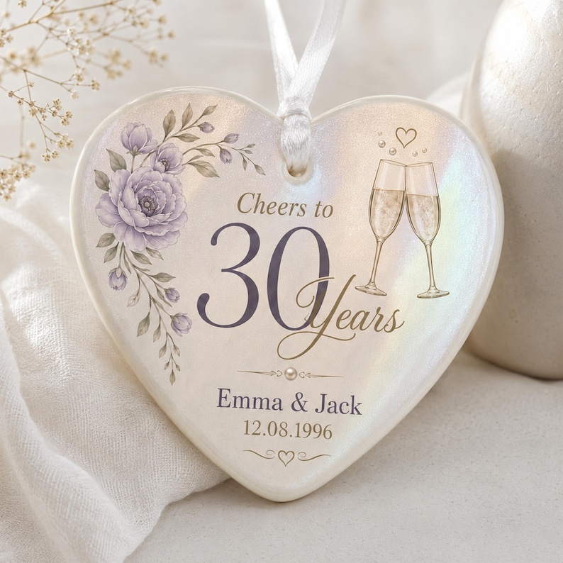 May include: A heart-shaped ceramic ornament with an iridescent finish, featuring a purple floral illustration, two champagne flutes, and the text Cheers to 30 Years, Emma & Jack, and 12.08.1996. A white ribbon is attached to the top for hanging.