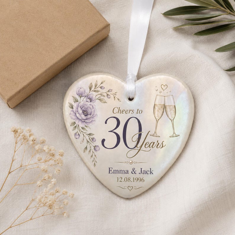 May include: A heart-shaped ceramic ornament with a white ribbon, featuring a purple floral illustration, two champagne flutes, and the text Cheers to 30 years, Emma & Jack, and 12.08.1996. The surface has an iridescent finish with a small heart icon.