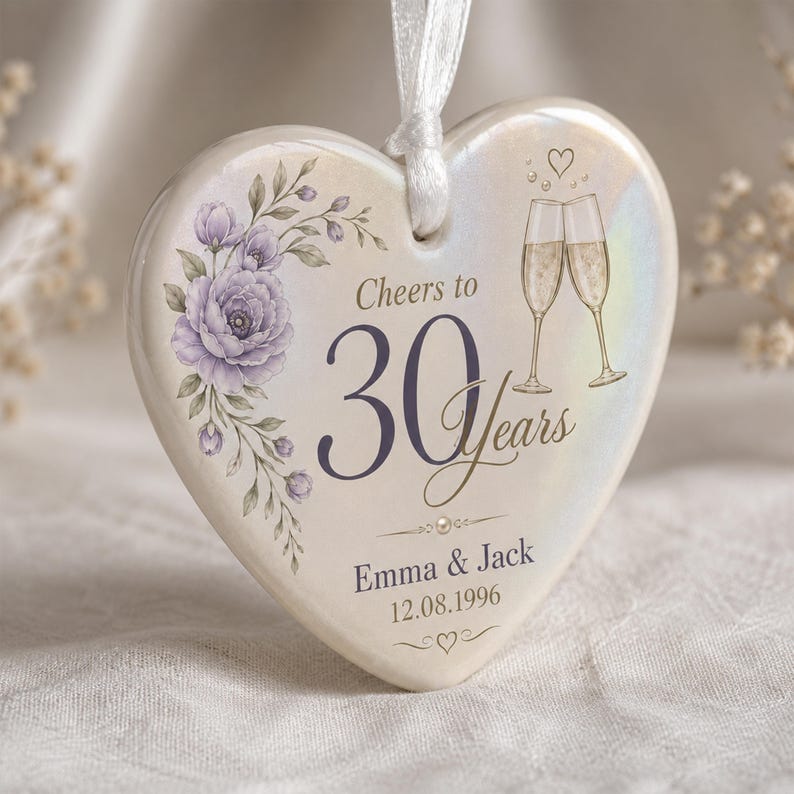 May include: A heart-shaped ceramic ornament with a white iridescent finish, featuring a purple floral illustration, two champagne flutes, and the text Cheers to 30 Years, Emma & Jack, and 12.08.1996. A white ribbon is attached to the top for hanging.
