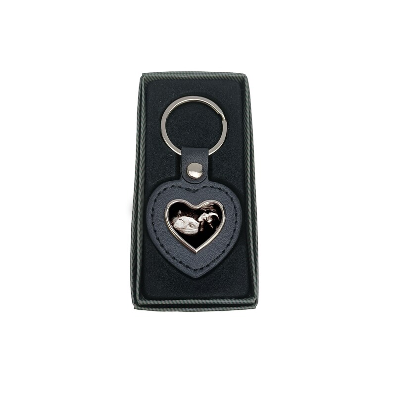 May include: A black leather heart-shaped keyring with a silver ring and a silver frame holding a black and white ultrasound image. The keyring is in a black velvet box.