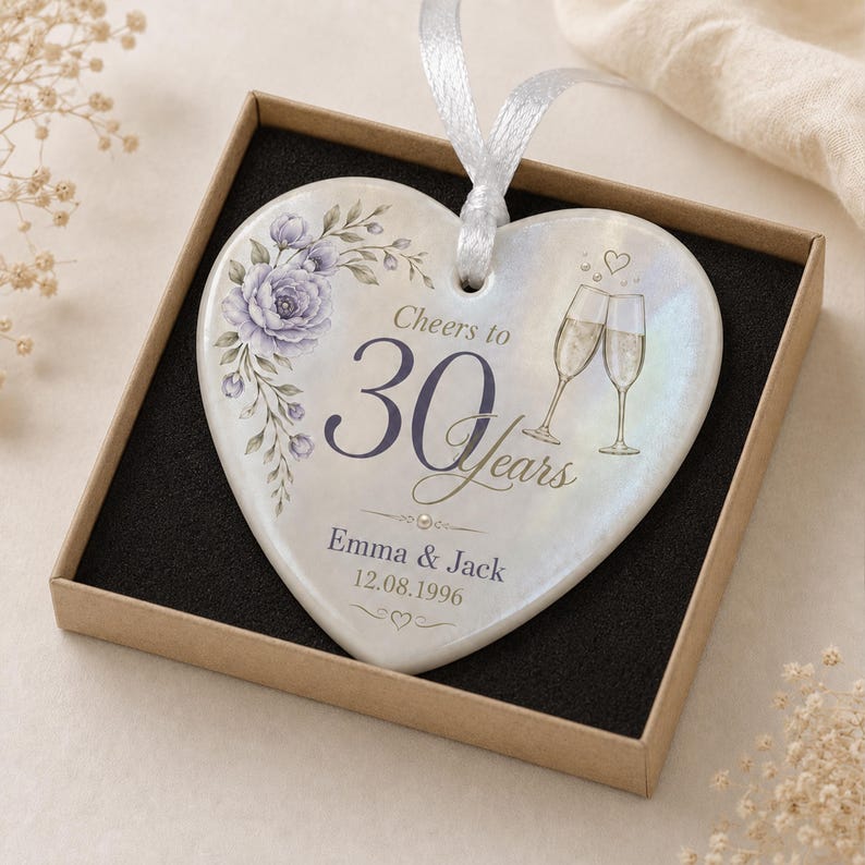 May include: A heart-shaped ceramic ornament with a pearlescent finish, featuring a purple floral illustration, two champagne flutes, and the text Cheers to 30 Years, Emma & Jack, 12.08.1996. It includes a white ribbon for hanging and a gift box.