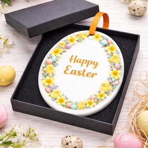 May include: Oval Easter ornament in a black gift box. The white ceramic ornament features the text "Happy Easter" in orange, surrounded by a floral border with yellow daffodils, chicks, and pastel eggs. An orange ribbon is attached for hanging.