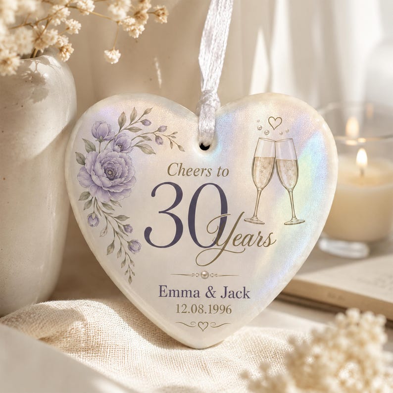 May include: A heart-shaped ceramic ornament with an iridescent finish, featuring a purple floral illustration, two champagne flutes, and the text Cheers to 30 Years, Emma & Jack, and 12.08.1996. A white ribbon is attached to the top for hanging.