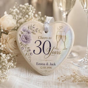May include: A heart-shaped ceramic ornament with a white iridescent finish, featuring a purple floral illustration, two champagne flutes, and the text Cheers to 30 Years, Emma & Jack, and 12.08.1996. It includes a white ribbon loop for hanging.