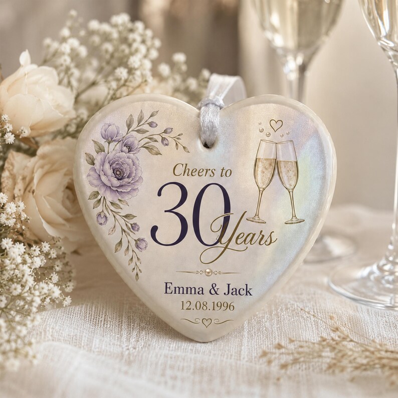 May include: A heart-shaped ceramic ornament with a white iridescent finish, featuring a purple floral illustration, two champagne flutes, and the text Cheers to 30 Years, Emma & Jack, and 12.08.1996. It includes a white ribbon loop for hanging.