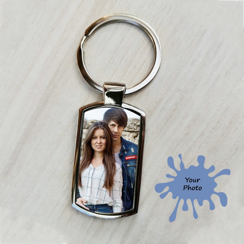 A silver rectangular metal keyring featuring a printed photo of a smiling couple. The keyring is placed on a light wooden surface with a blue paint splash graphic next to it reading "Your Photo."