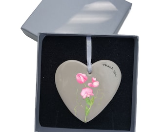 May include: A gray ceramic heart-shaped ornament with a pink sweet pea flower design and the text "Thank you" on the right side. The ornament is in a gray gift box with a black interior.