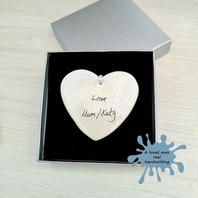 May include: A white ceramic heart ornament with iridescent detailing. The heart has the words "Love Mum/Katy" written on it in black ink.