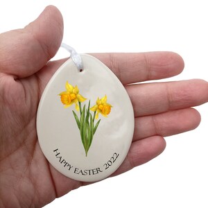 Pode incluir: Ornamento de cer&acirc;mica branca em forma de ovo com um desenho de flores de narciso amarelo e o texto "Happy Easter 2022".