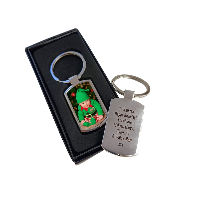 A silver photo keyring in a black gift box with a printed image of a baby in a green Christmas elf outfit. Next to it is the back of another keyring engraved with a birthday message addressed to Kathryn from Melissa, Garry, Chloe, AJ, and Willow-Rose.