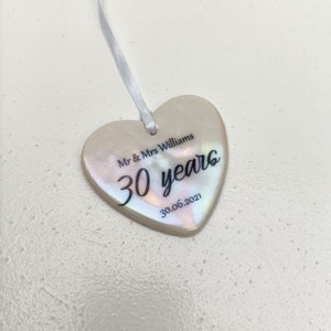 May include: A white heart-shaped ceramic ornament with iridescent detailing. The ornament has black text that reads "Mr & Mrs Williams 30 years 30.06.2021".