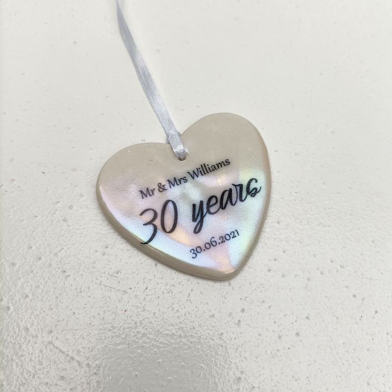 May include: A white heart-shaped ceramic ornament with iridescent detailing. The ornament has black text that reads "Mr & Mrs Williams 30 years 30.06.2021".