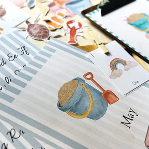 May include: A collection of watercolour illustrations with a beach theme, including a sand pail, shovel, and rainbow. The image features a blue and white striped background with the word "May" and "Morning Menu". There are also alphabet letters and sea creature stickers.