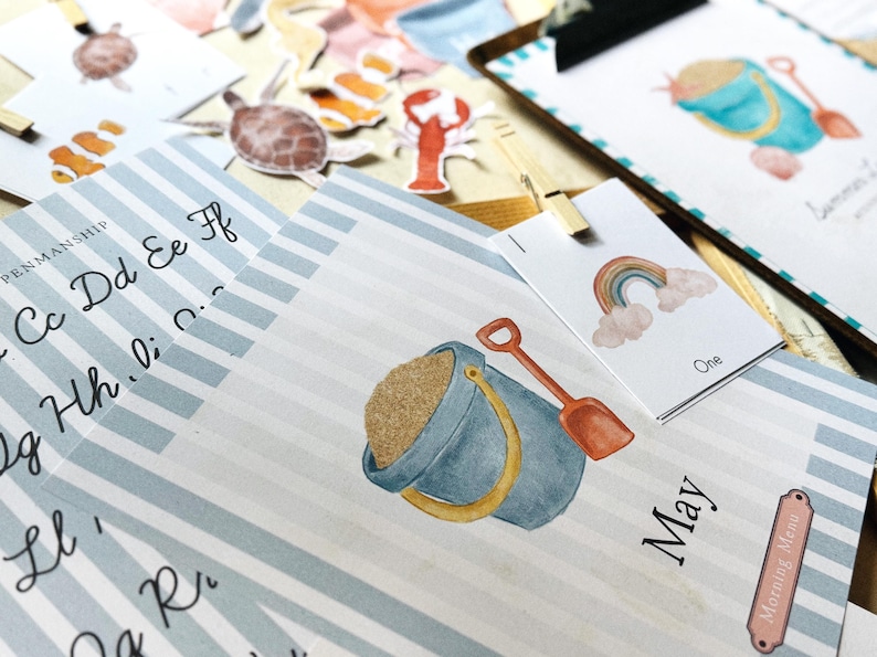 May include: A collection of watercolour illustrations with a beach theme, including a sand pail, shovel, and rainbow. The image features a blue and white striped background with the word "May" and "Morning Menu". There are also alphabet letters and sea creature stickers.