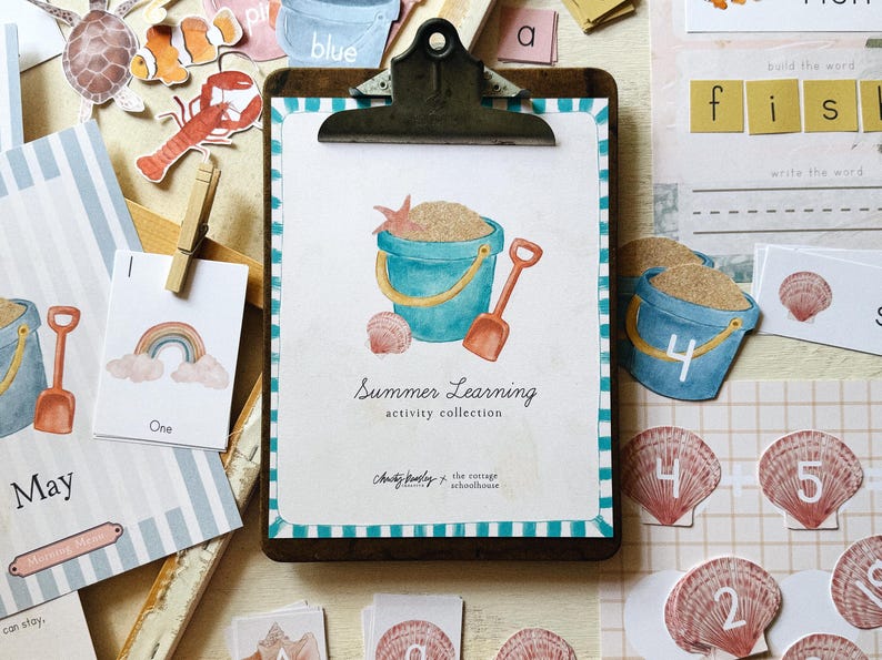 May include: A wooden clipboard featuring a watercolour illustration of a sand pail, shovel, and seashells. The text "Summer Learning activity collection" is displayed. Additional cards with numbers, letters, and sea creatures are scattered around.
