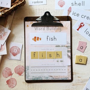 May include: A wooden clipboard with the words "Word Building" and "fish" printed on it. The image includes word cards, letter tiles, and illustrations of a clownfish, shell, ice cream, and lobster. The word "fish" is spelled out with letter tiles.