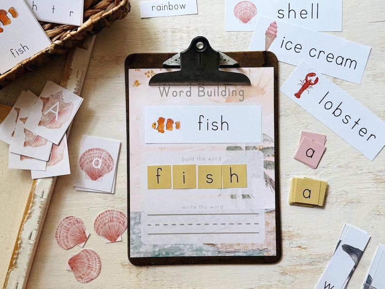 May include: A wooden clipboard with the words "Word Building" and "fish" printed on it. The image includes word cards, letter tiles, and illustrations of a clownfish, shell, ice cream, and lobster. The word "fish" is spelled out with letter tiles.