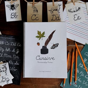 May include: An assortment of cursive handwriting resources. Includes a spiral-bound book titled "Cursive Penmanship Primer" with illustrations of a feather, inkwell, leaf, and moth. Also features alphabet cards, a chalkboard, and pencils.