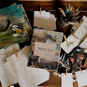 May include: An overhead shot of a wooden table with books, maps, and art supplies. The book "The Discovery of the Americas" is open, and the book "Woven: History & Geography Series" is visible. There are also paintbrushes, paint tubes, and small cards.