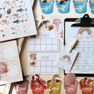 May include: Educational resources with a seaside theme. Includes number cards, a clipboard with number writing practice, and various sea creature cutouts. The colour palette features blues, pinks, and yellows. Designed for early childhood education.
