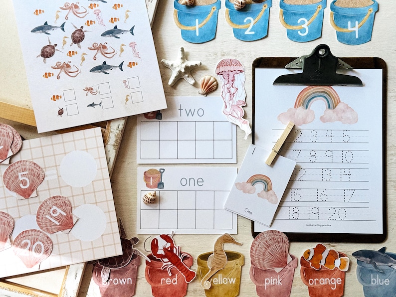 May include: Educational resources with a seaside theme. Includes number cards, a clipboard with number writing practice, and various sea creature cutouts. The colour palette features blues, pinks, and yellows. Designed for early childhood education.